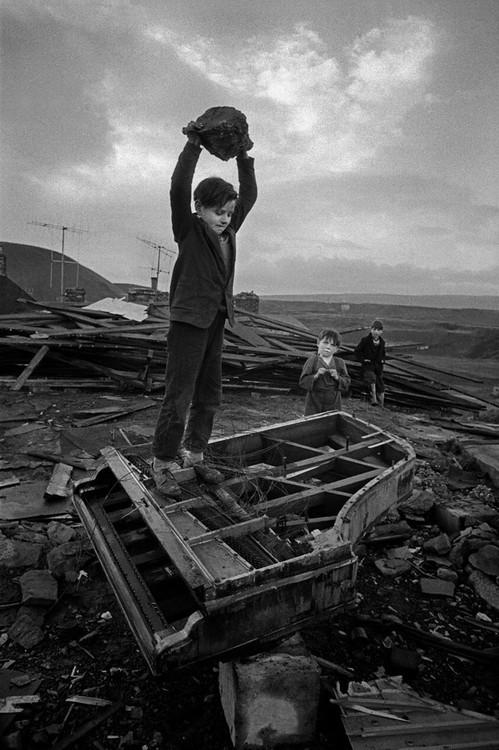 Boy Destroying Piano with a large rock under a leaden sky.